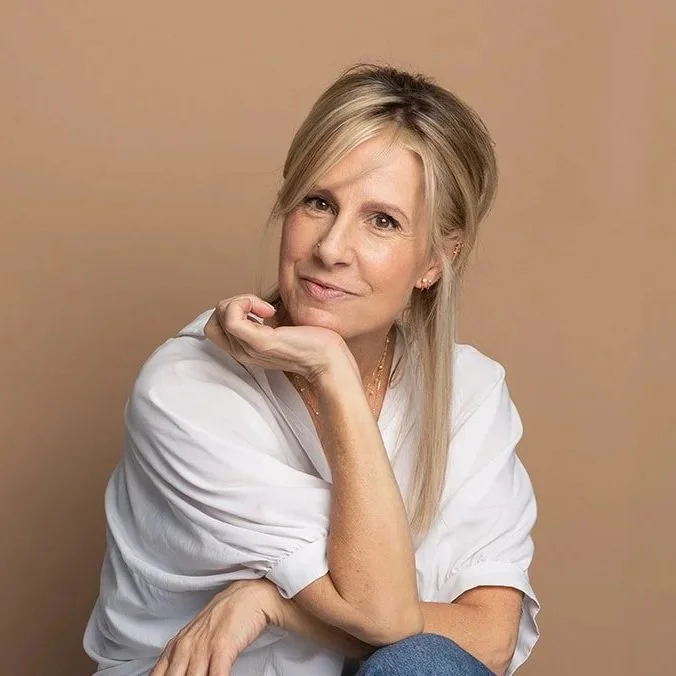 A woman with blonde hair in a casual white shirt, sitting against a light beige background, resting her chin on her hand and smiling softly at the camera.