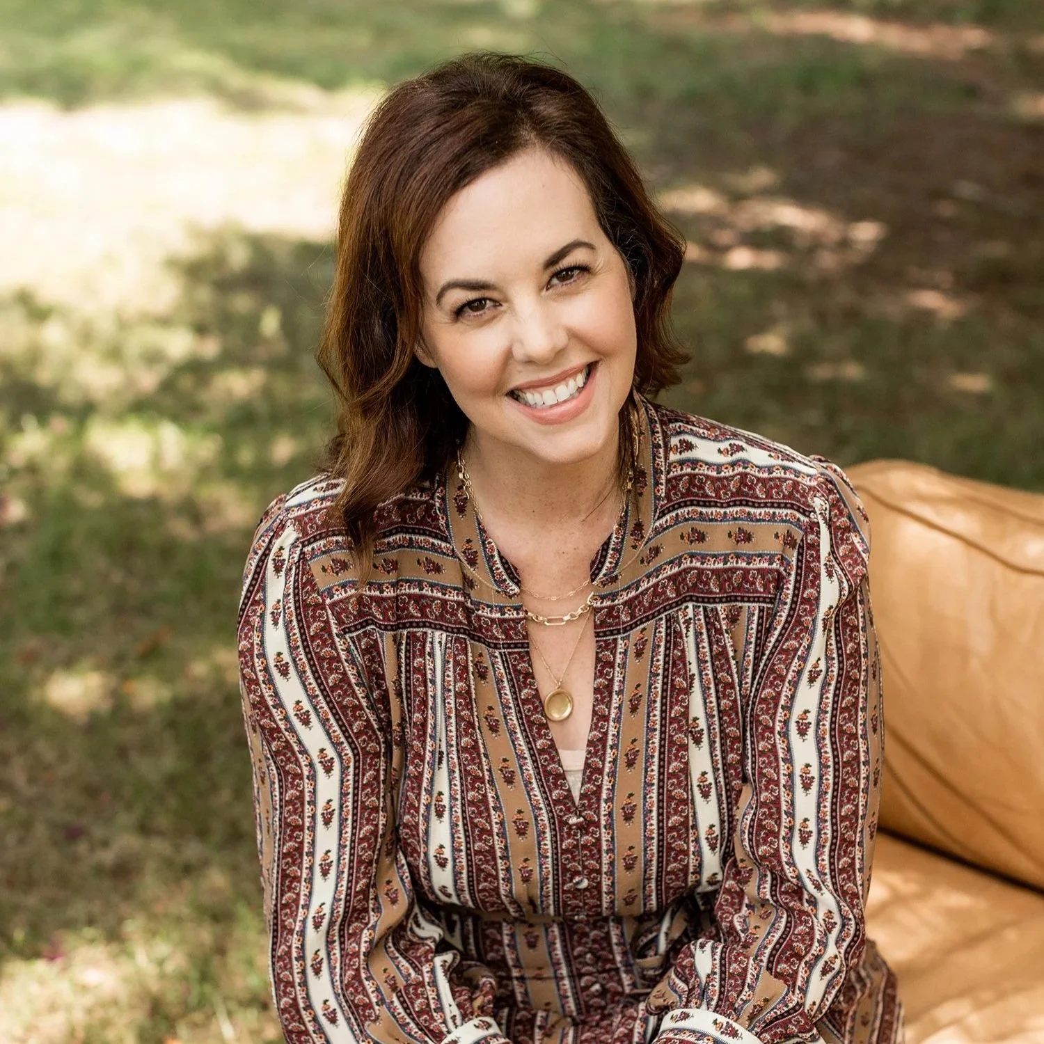 A woman with shoulder-length dark brown hair smiling outdoors, wearing a patterned blouse with red, brown, and white stripes and floral designs, and layered gold necklaces.