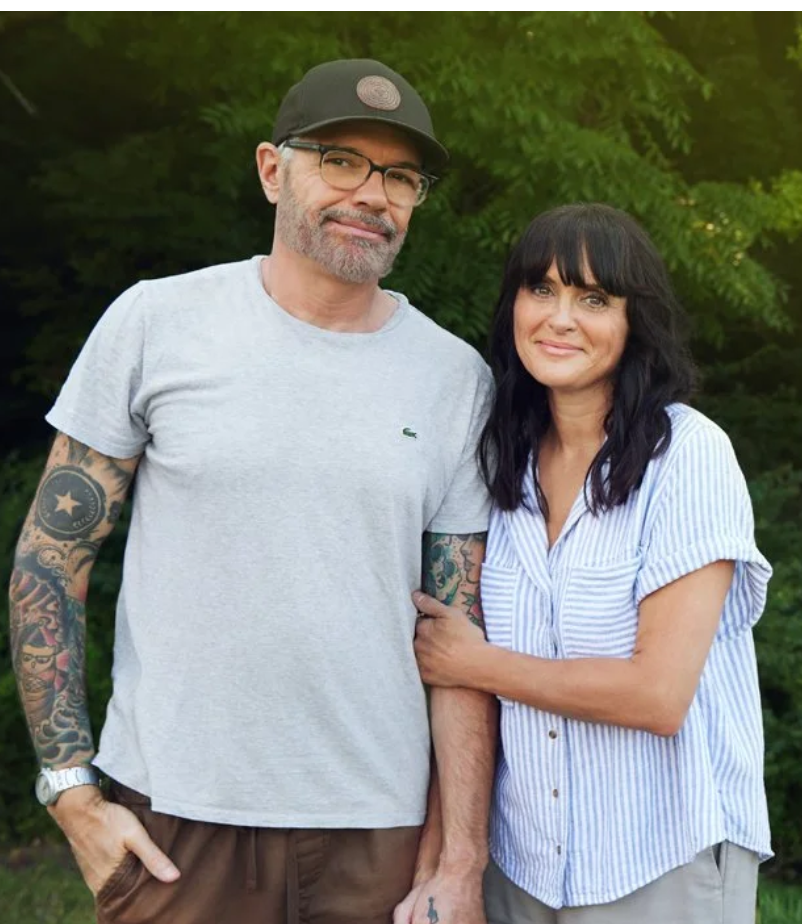 A man and woman stand outdoors in front of green trees, smiling gently at the camera. The man wears glasses, a black cap, a light gray T-shirt, and has tattooed arms. The woman has black hair, wears a blue and white striped short-sleeve blouse, and has her arm around the man.