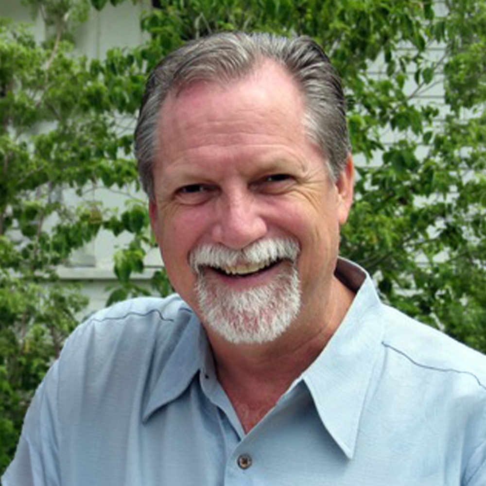 A smiling middle-aged man with gray hair and a beard, wearing a light blue collared shirt, standing outdoors with green foliage in the background.