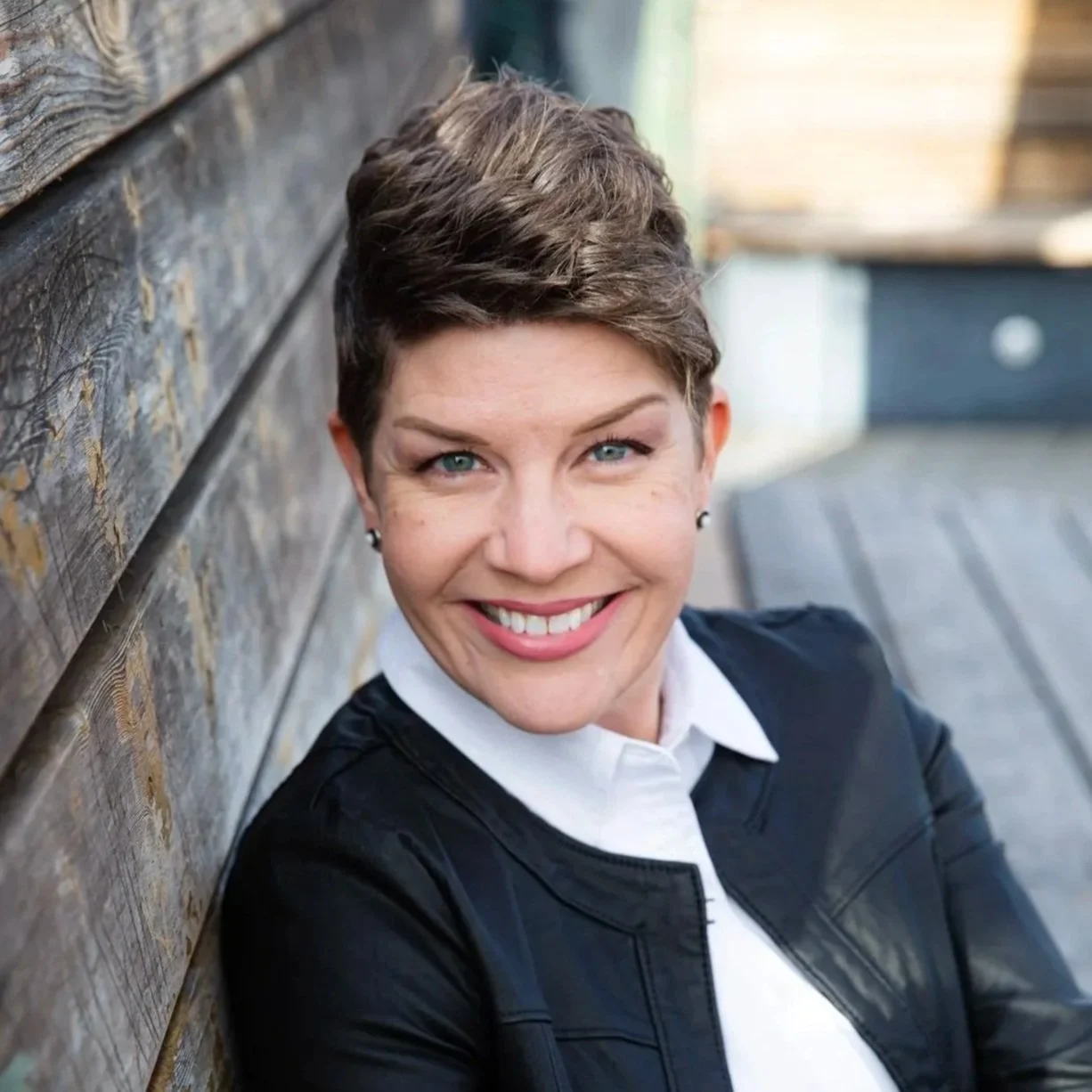 Close-up of a smiling woman with short brown hair, wearing a white shirt and black leather jacket, sitting outdoors against a wooden wall.
