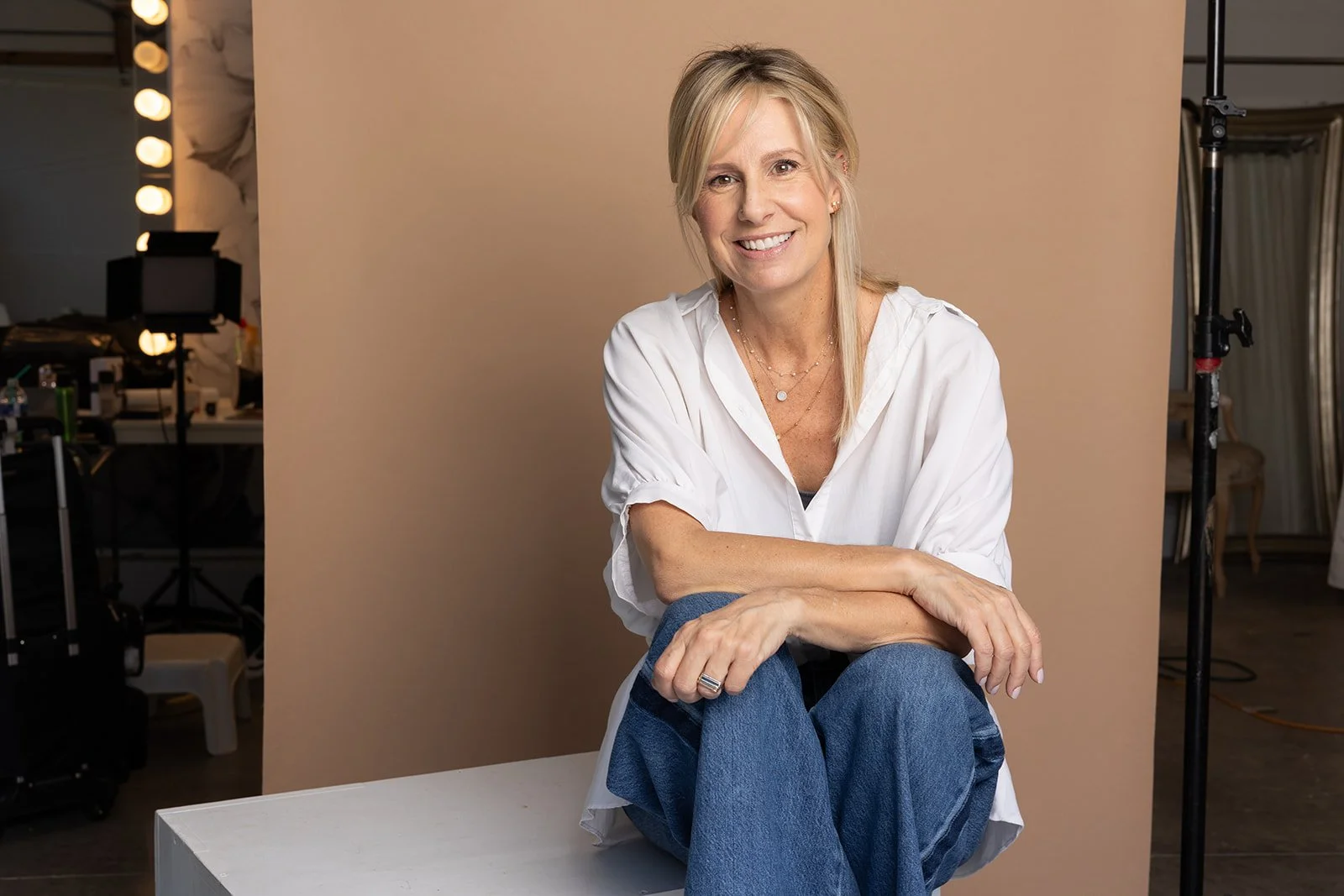 Smiling blonde woman sitting on a table with legs crossed, in a photography studio with beige background, wearing a white shirt and blue jeans.