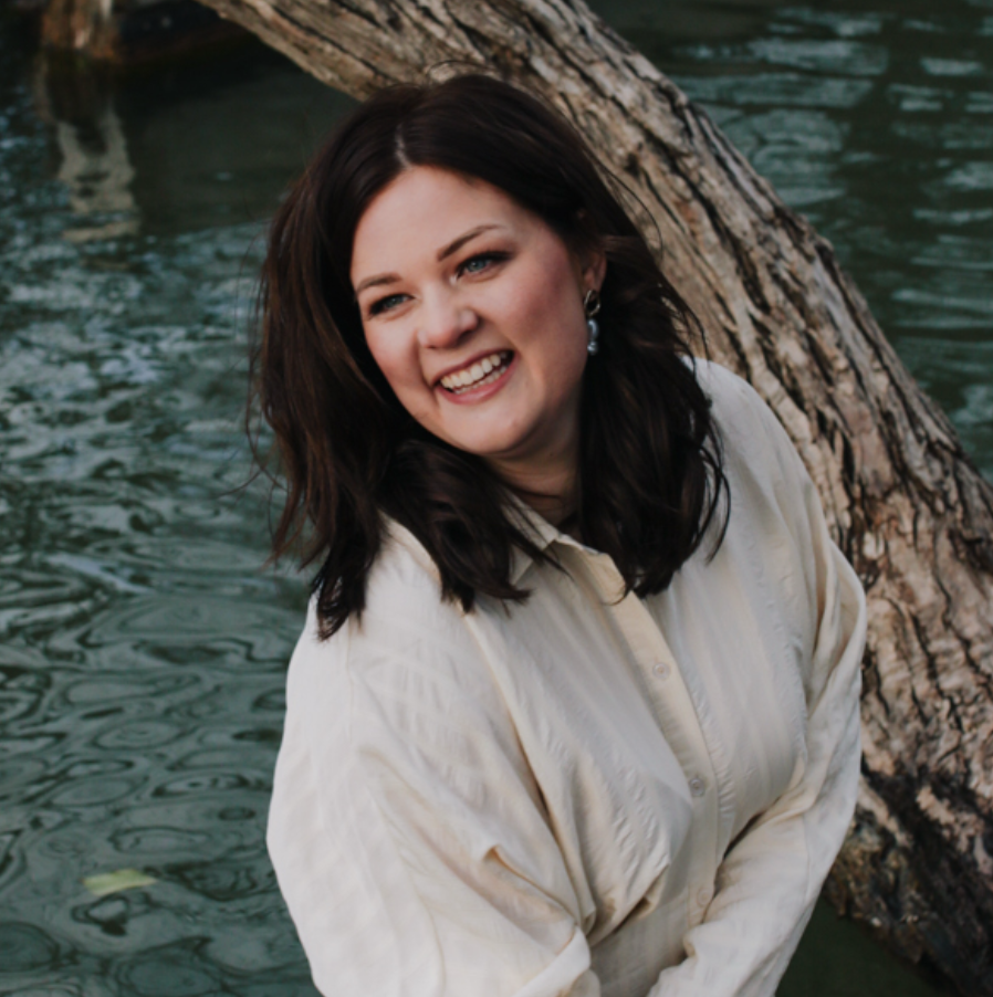 Woman with dark brown hair smiling near a tree trunk and water.