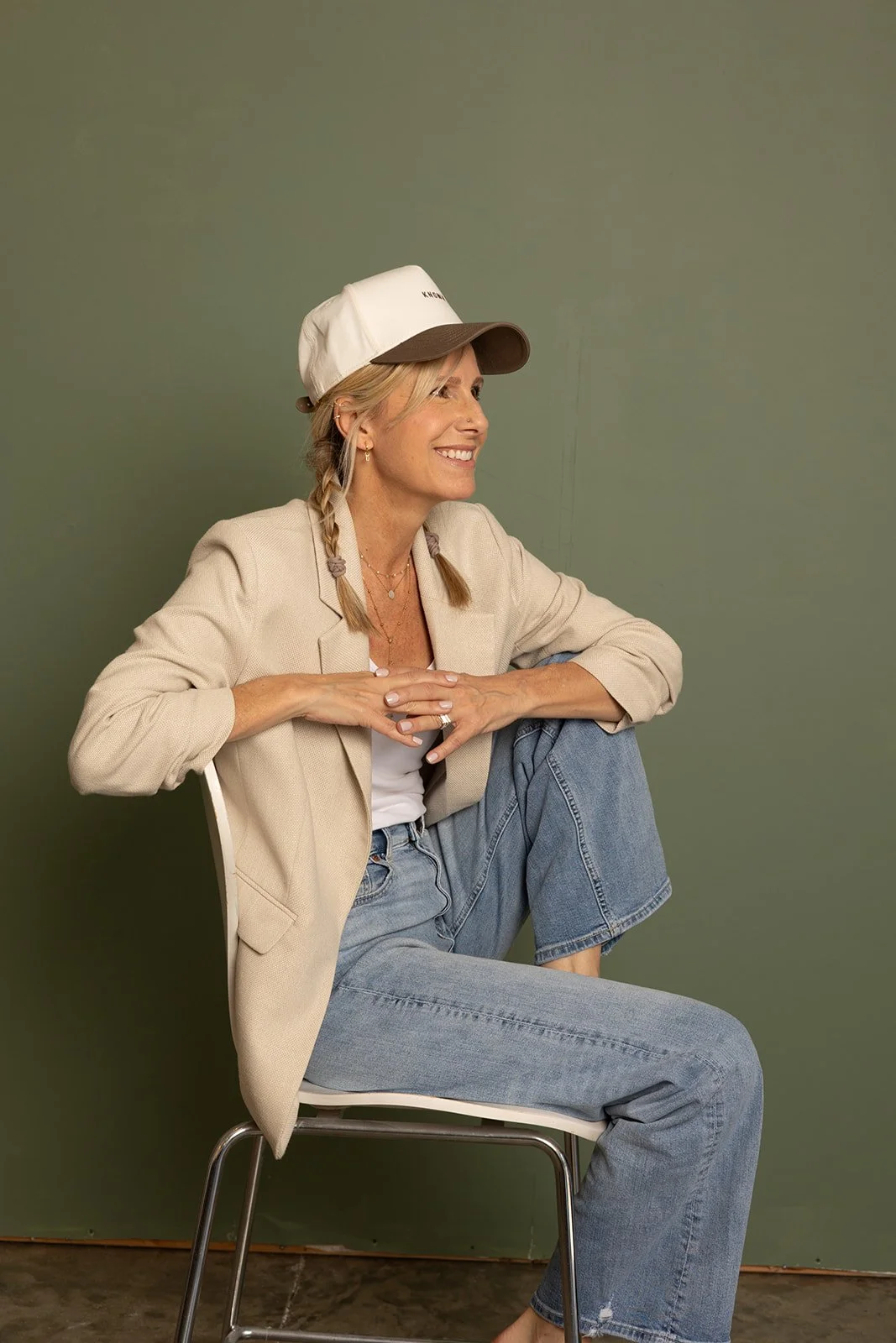 A woman with blonde hair in braids, wearing a beige blazer, white t-shirt, light-wash jeans, and a beach cap, sitting on a chair with one leg up, smiling, against a green wall.