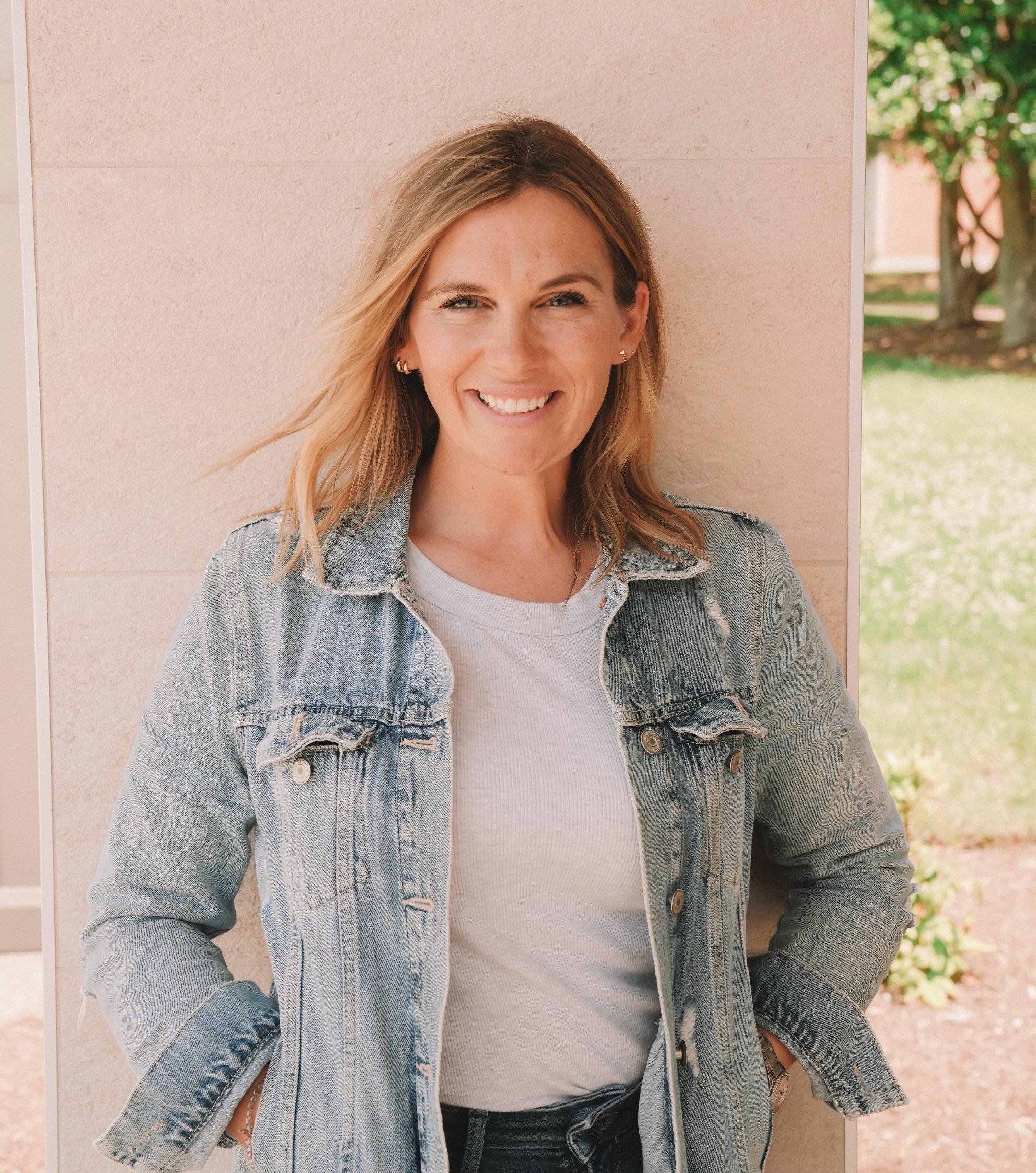 Portrait of a smiling woman with blond hair, wearing a denim jacket over a white shirt, standing outdoors near a peach-colored wall with green trees in the background.