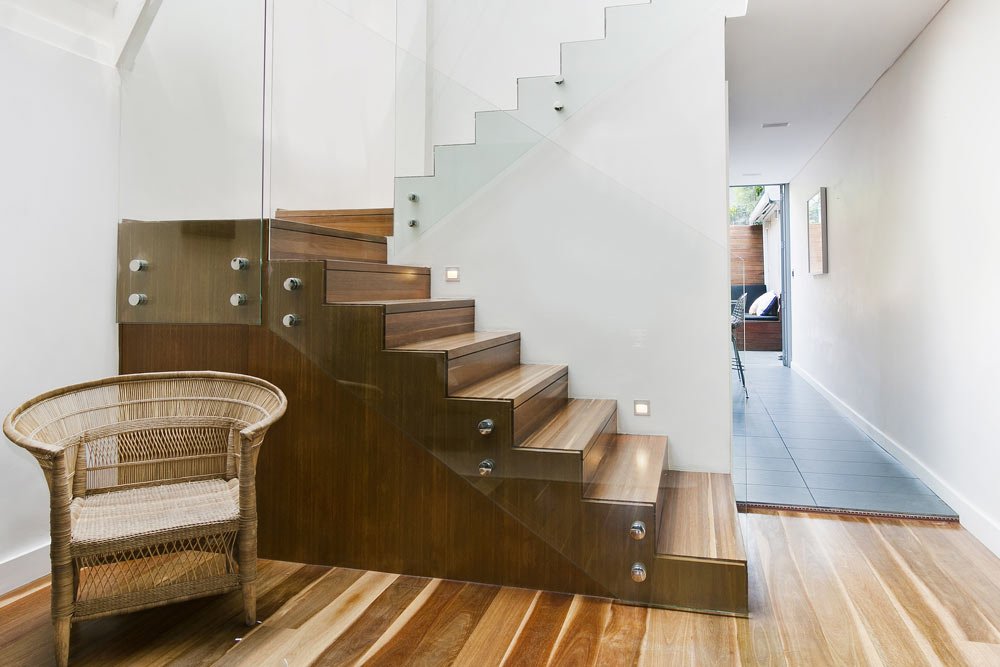 Modern interior staircase with wooden steps, glass railing, small wall lights, and a wicker chair in a bright hallway.