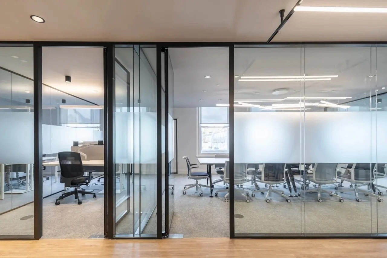 Modern glass-walled conference room with office chairs around a table and a window in the background.