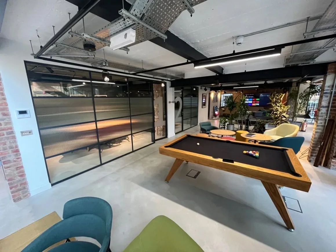 Modern lounge with a pool table, seating area, plants, and a glass wall dividing rooms.