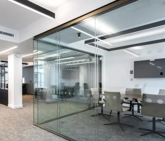 Modern office conference room with glass walls, a large TV screen, and six beige office chairs around a rectangular table.