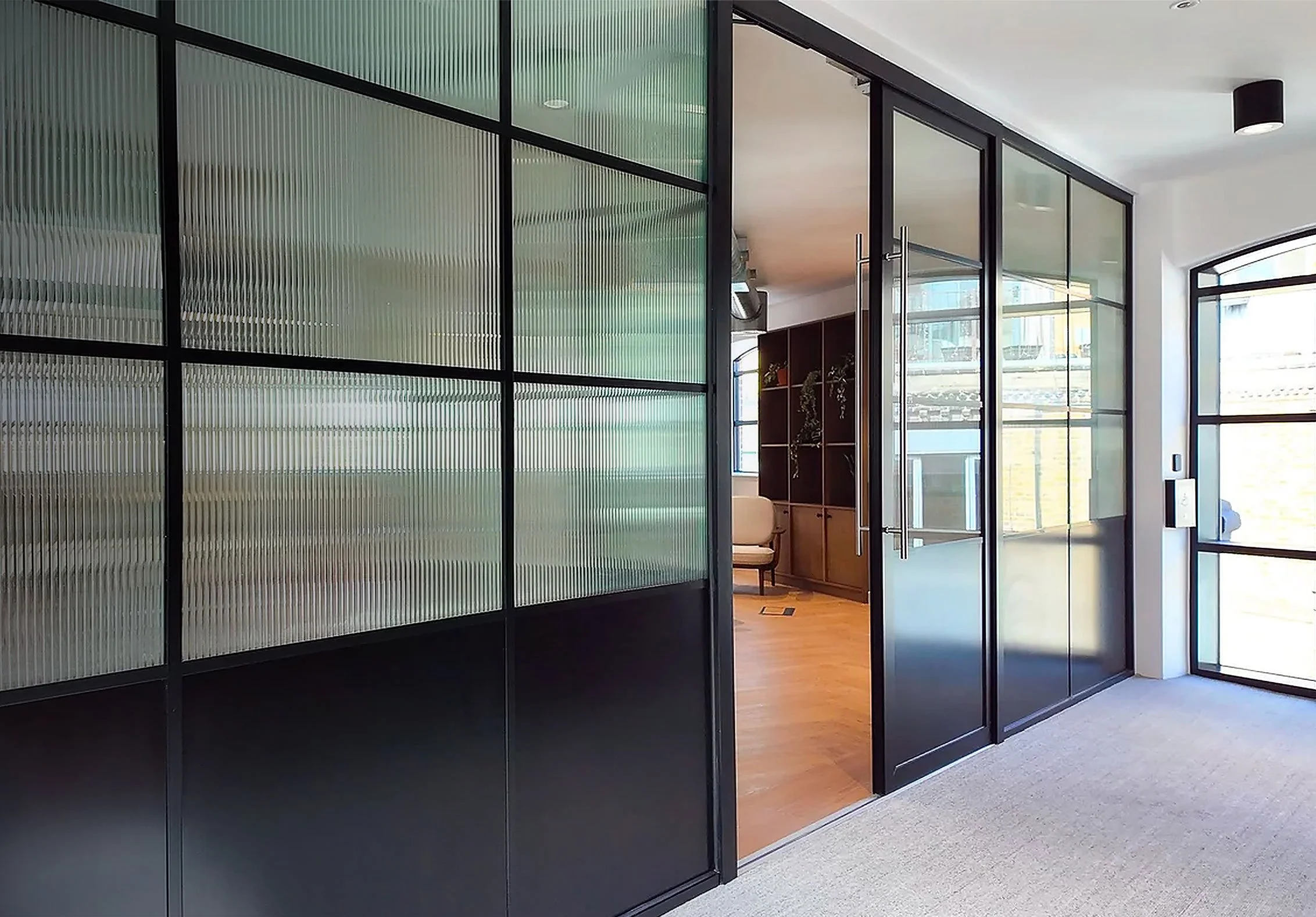 Modern interior with black-framed glass partition walls, wooden flooring, white walls, and a large window letting in natural light.