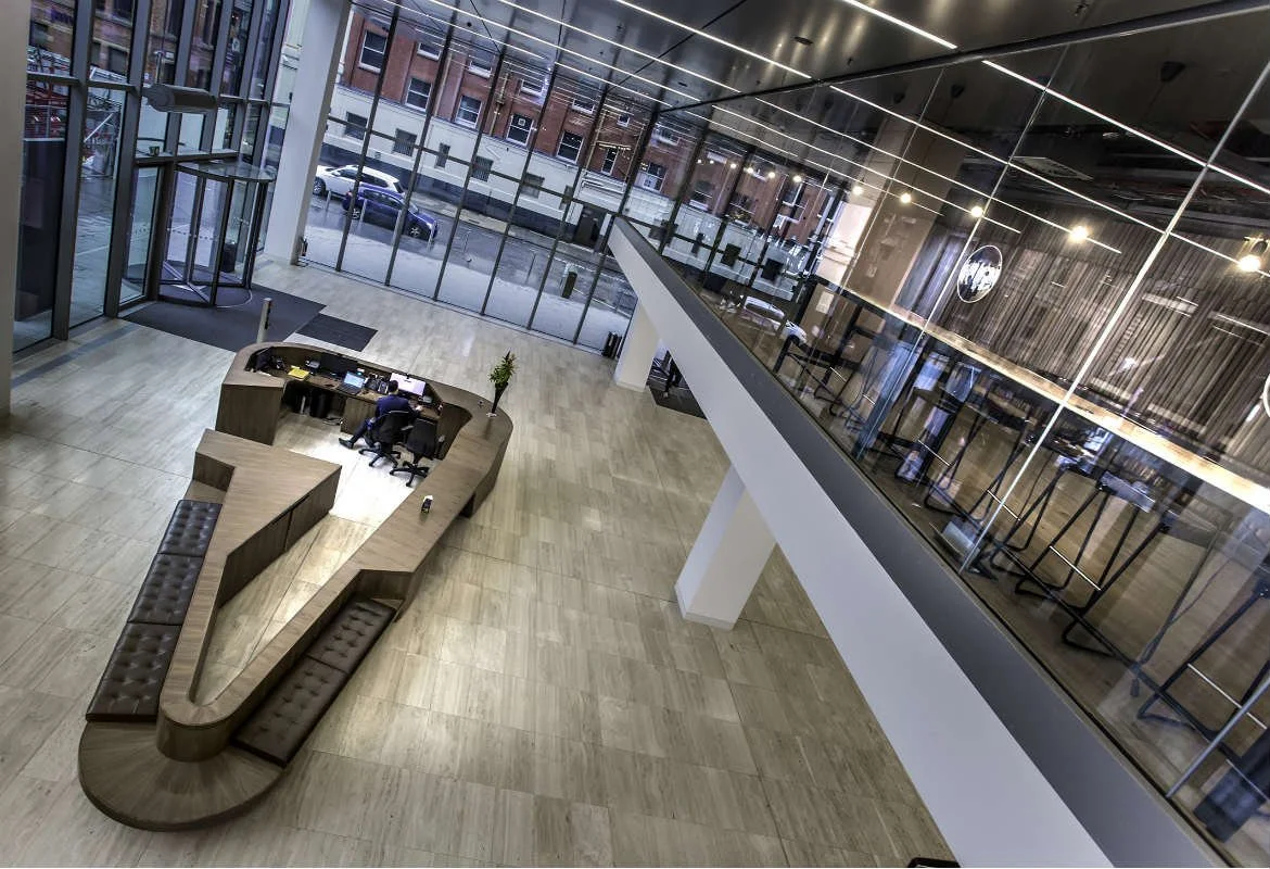 Interior view of a modern office lobby with a curved reception desk, a man working at the desk, floor-to-ceiling glass windows, and a second-floor seating area with barstools.