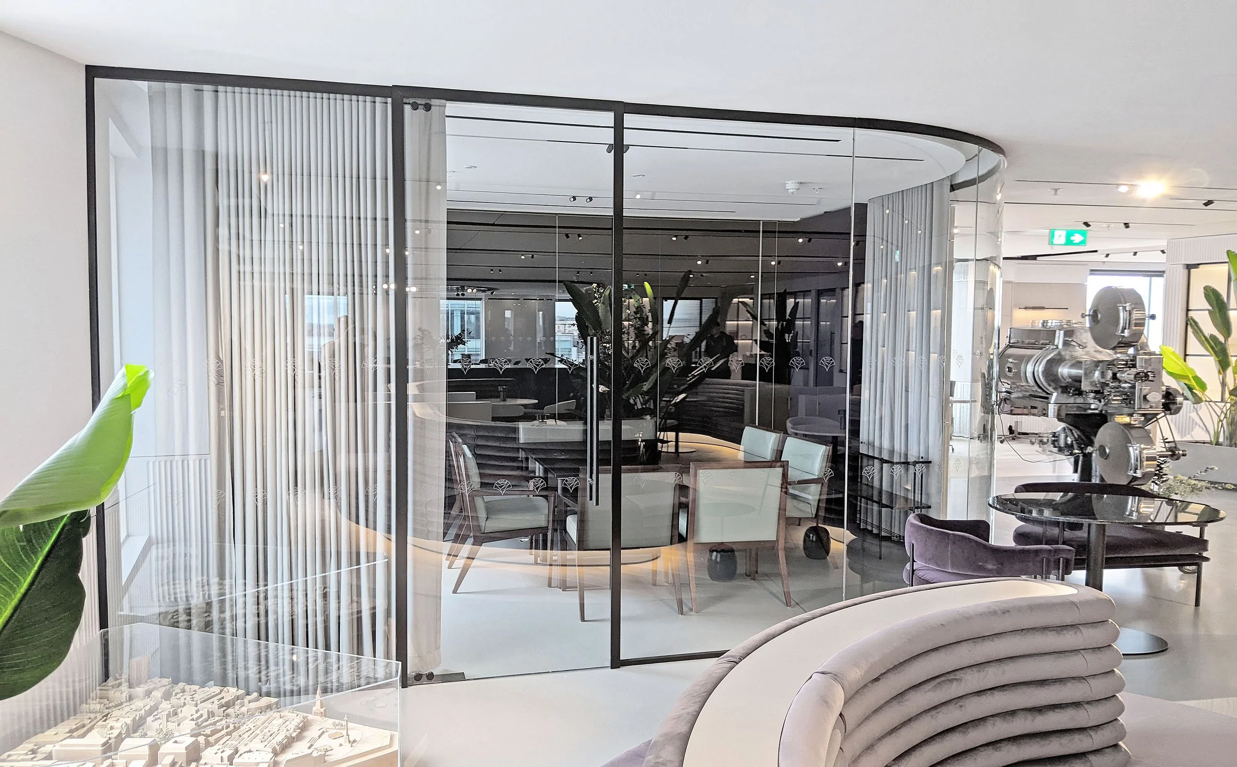 Interior view of a modern building with glass walls, white curtains, and seating areas, including a curved velvet sofa. There is a large piece of machinery on the right side and a model of a city or development plan at the bottom left.