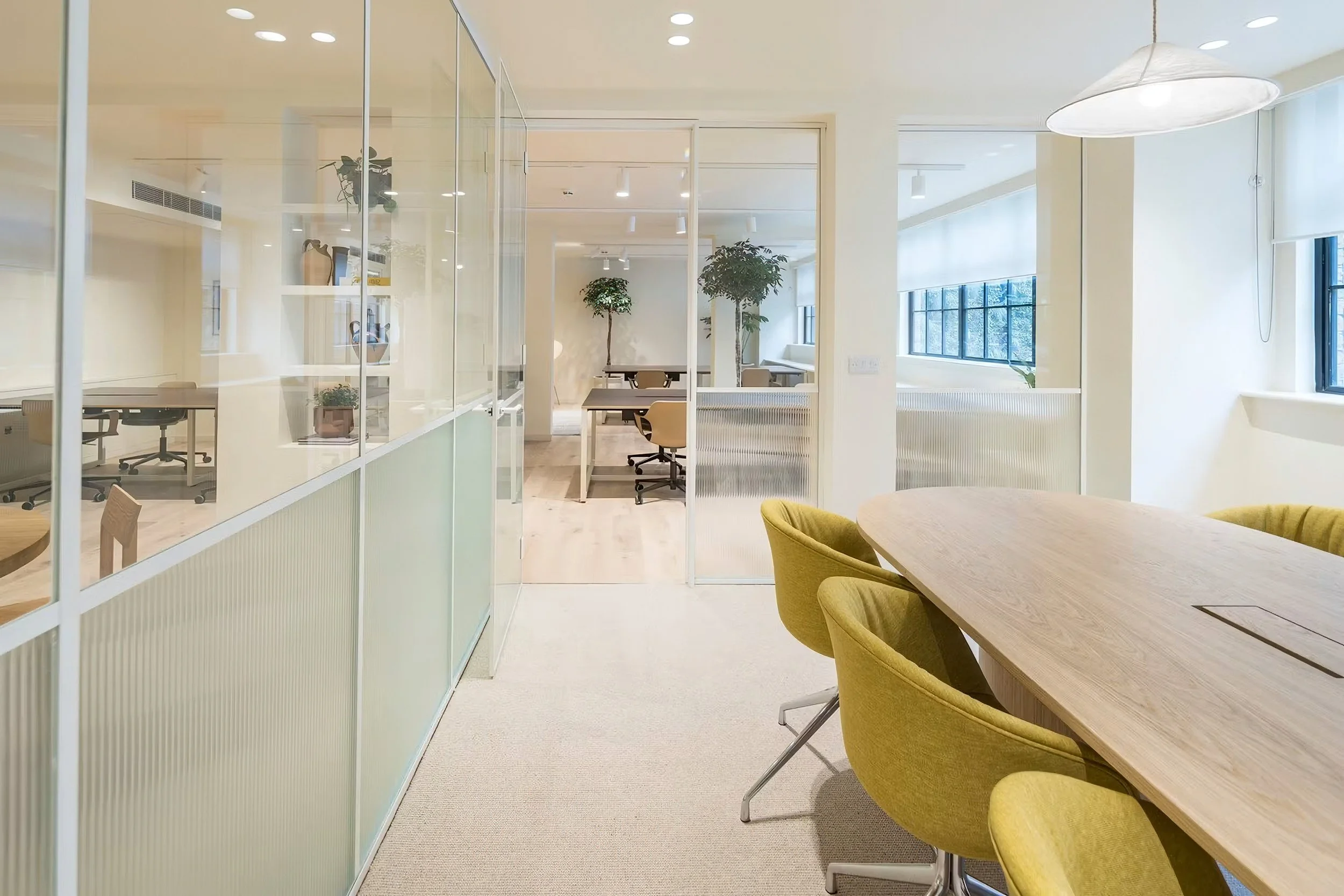 Office space with glass partitions, wooden tables, yellow chairs, potted plants, large windows with blinds, and a ceiling pendant light.