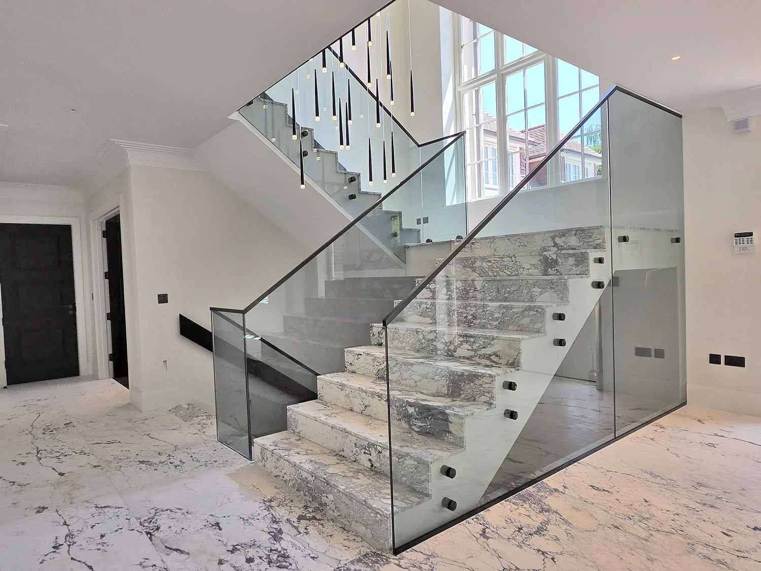 Modern interior staircase with marble steps, glass panels, and black handrails, in a well-lit room with large windows and white walls.