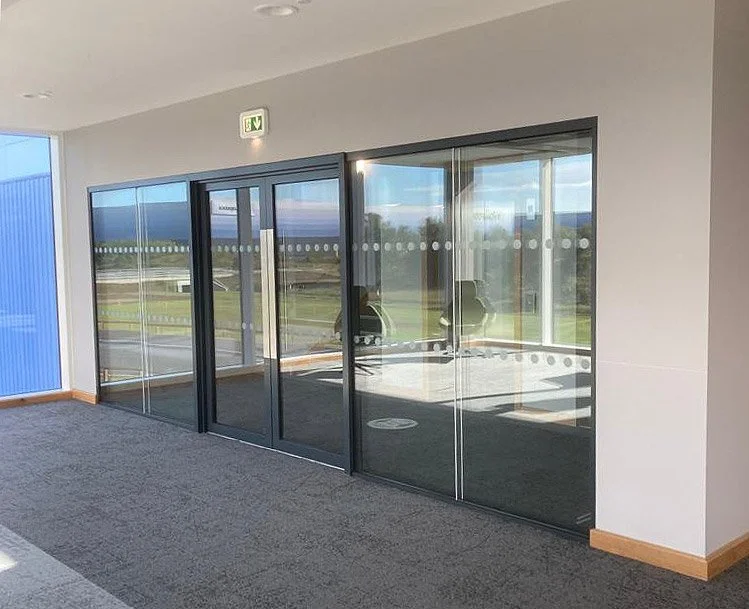 Interior view of a modern office with sliding glass doors and a carpeted floor, with an external view of a parking lot and green landscape