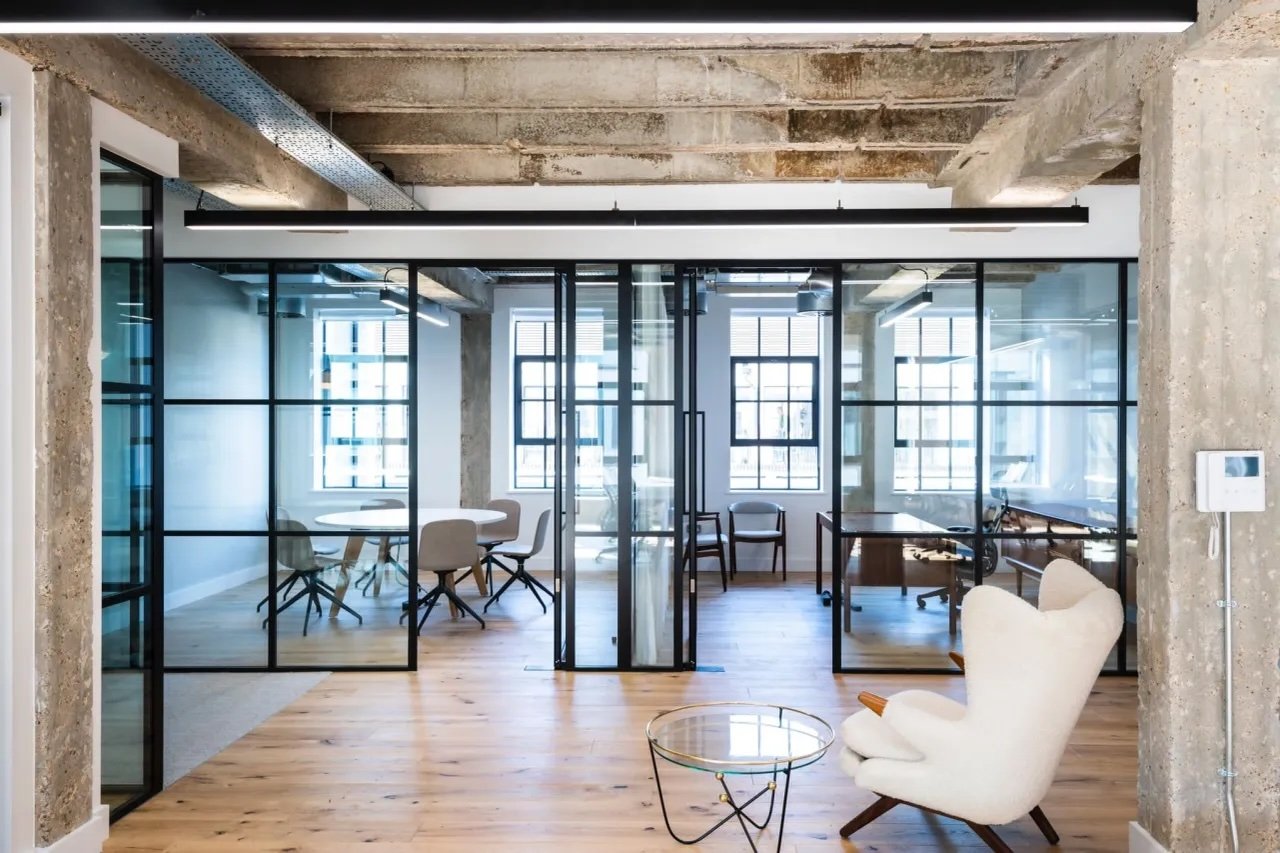 Modern office space with glass partitions, wooden floors, and furniture including a white armchair, round table, and desks. Large windows allow natural light in, and industrial-style exposed concrete beams are on the ceiling.