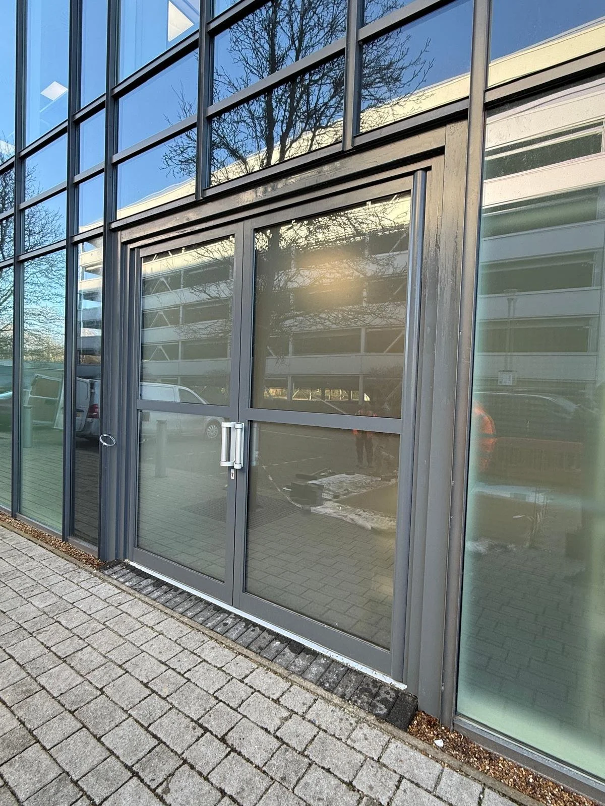 Exterior of a modern building with large glass windows and a glass door, reflecting trees and parked cars, and new brick paving outside.
