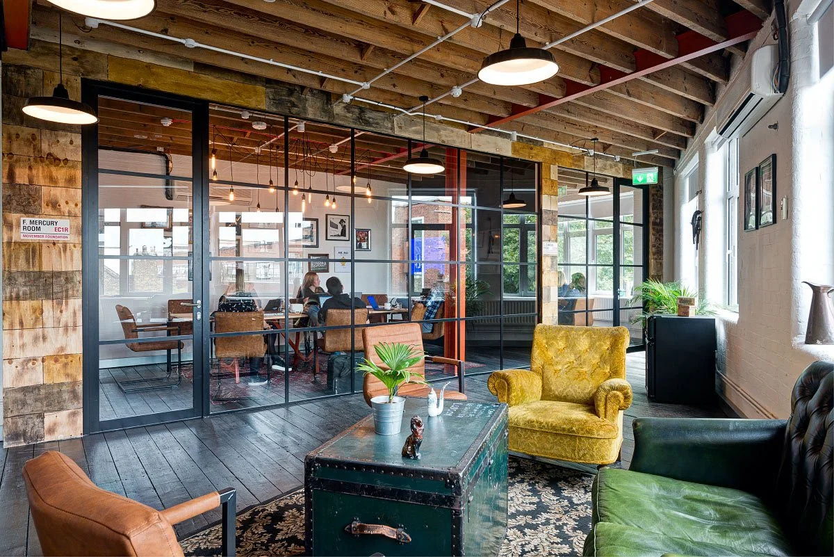 Cozy coworking space with glass walls, wooden ceiling, and vintage furniture. People are working at tables behind glass doors, while a seating area in the foreground includes a green velvet sofa, a yellow armchair, and a coffee table with plants and 