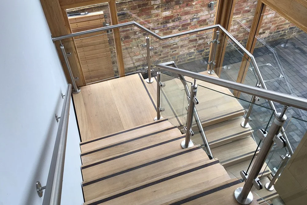 Wooden staircase with glass and metal railings viewed from above, leading down to a lower level with brick wall and glass window.