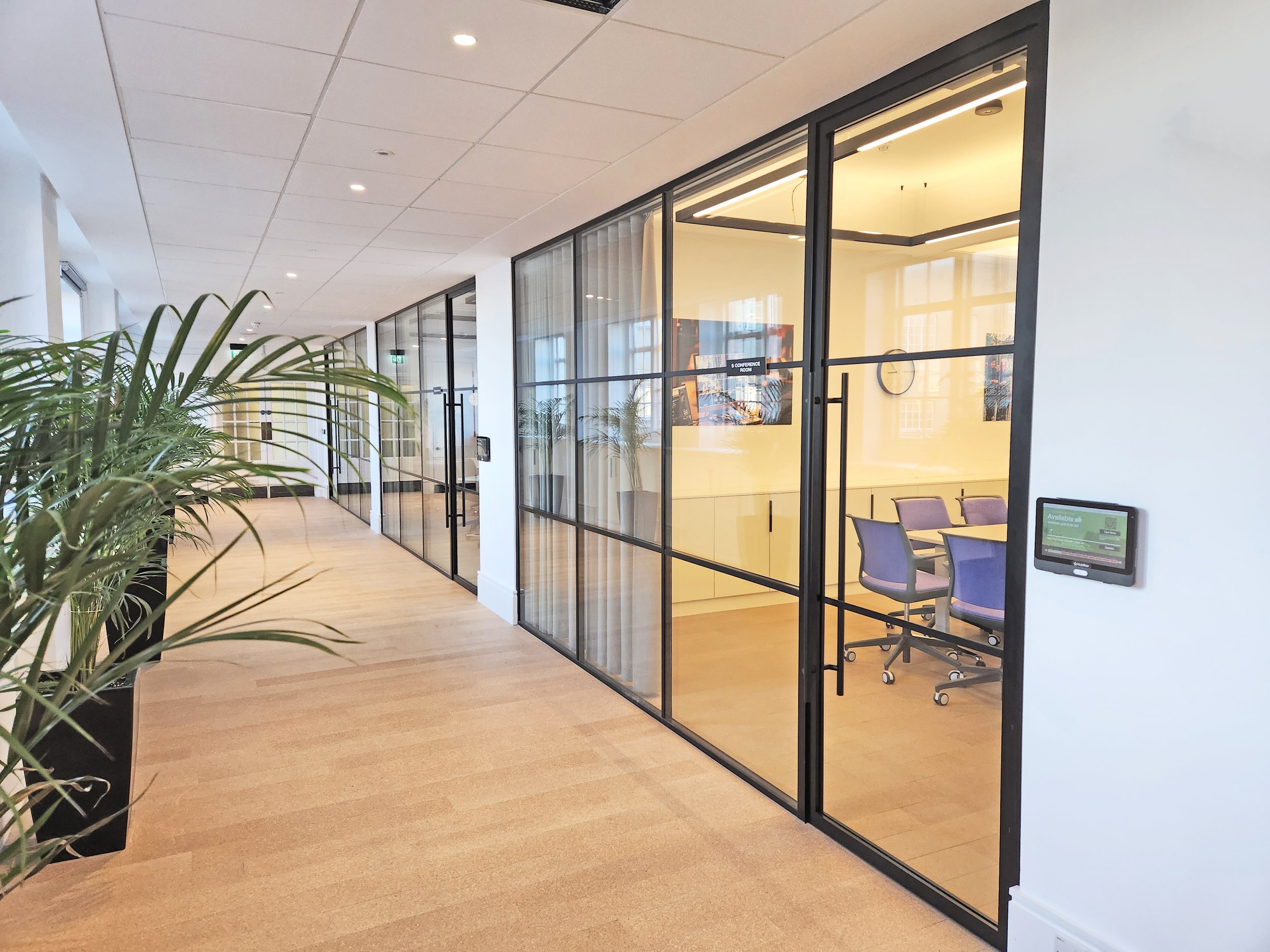 Office interior with conference rooms, glass walls, and plants along a hallway with beige carpet and white ceiling.