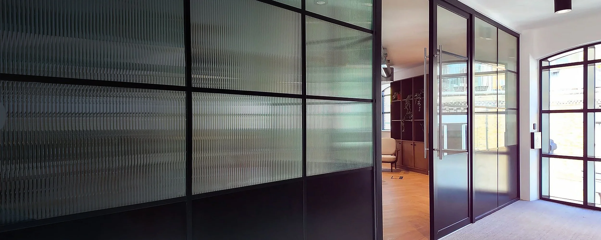 Modern office interior with glass walls, wooden flooring, a bookshelf, and large windows allowing natural light.