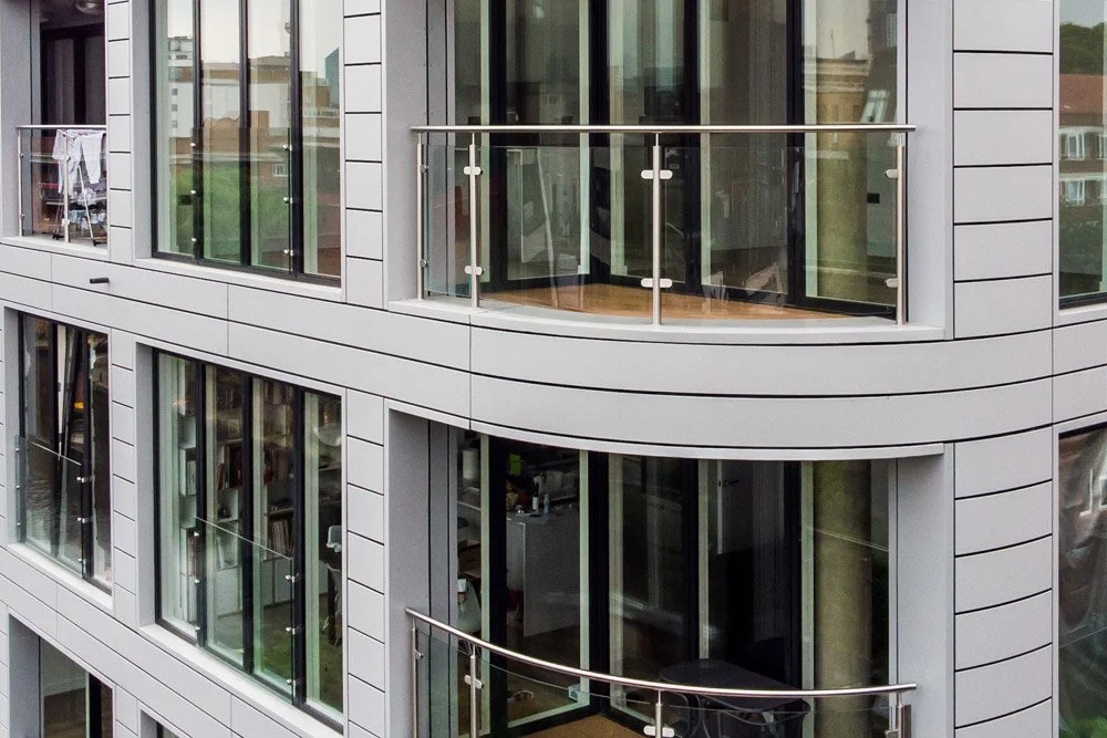 Exterior view of a modern residential apartment building with glass sliding doors, balconies with metal railings, and visible interior rooms.