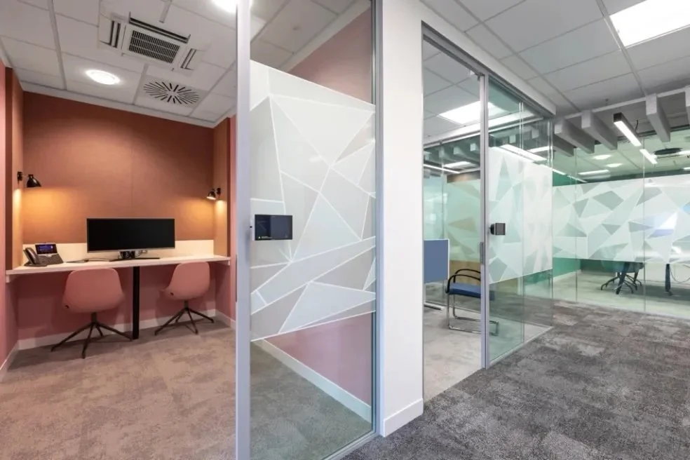 Two office rooms separated by glass walls in a modern office space. The room on the left has a pink wall, a desk with a computer monitor, a phone, and two pink chairs. The room on the right has glass walls with geometric patterns, chairs, and a confe