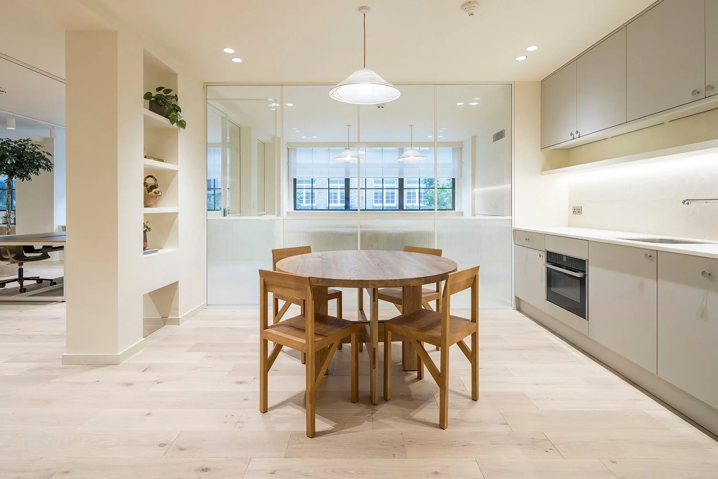 Minimalist kitchen and dining area with round wooden table and four chairs, white cabinetry, large windows with blinds, and pendant lighting fixtures.