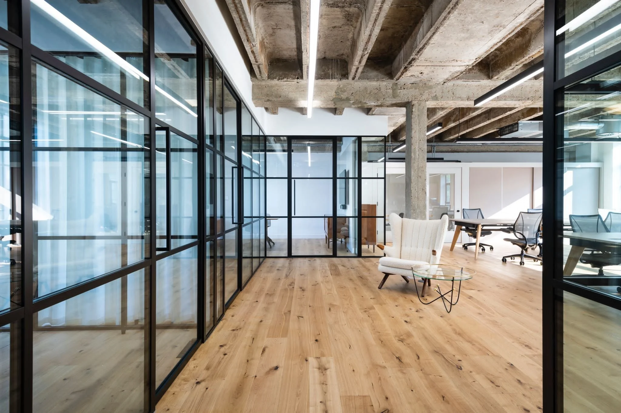 Modern office space with glass partitions, exposed concrete ceiling beams, wooden flooring, and minimalist furnishings.
