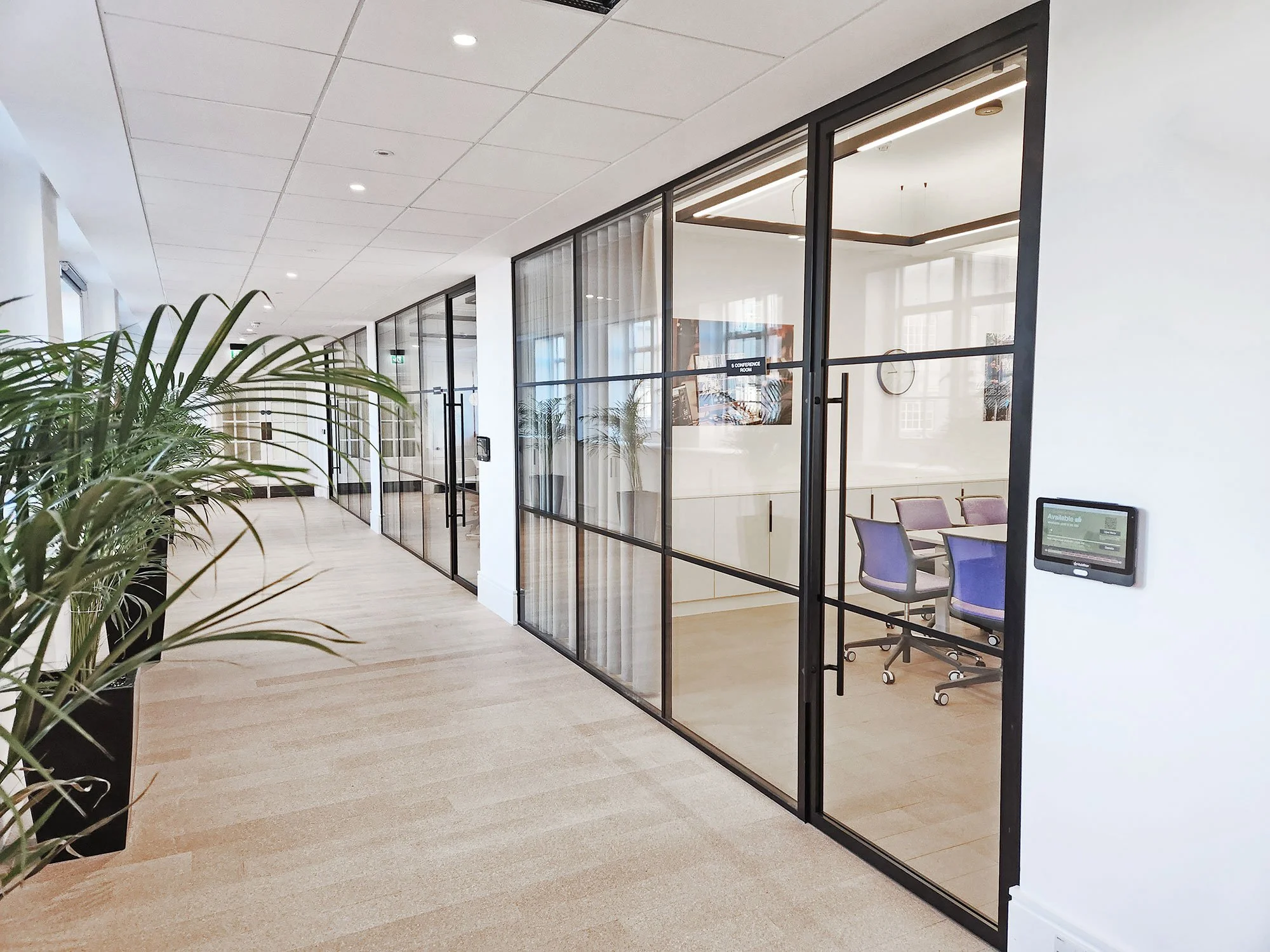 Modern office conference room with glass walls, purple chairs, a clock on the wall, and indoor plants in the hallway.