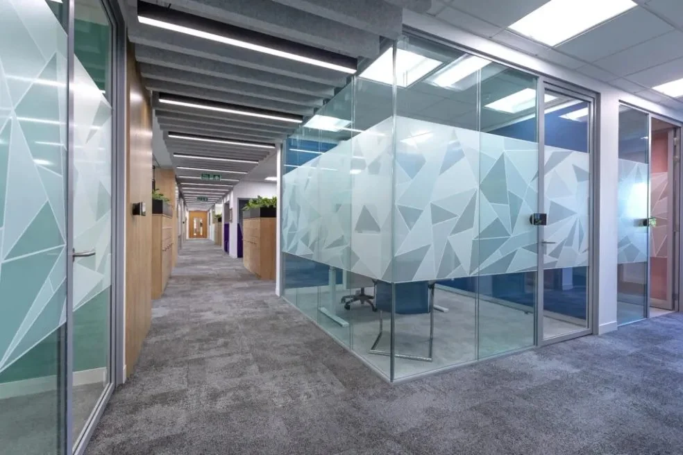 Modern office corridor with glass-walled meeting rooms, gray carpet, and ceiling with linear lighting.