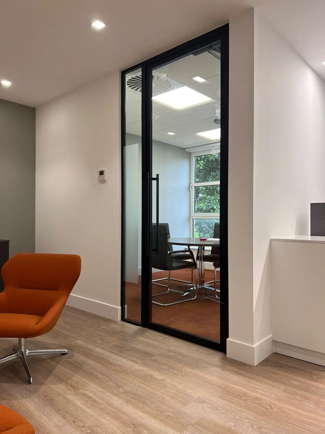 Modern office space with glass door leading to conference room, orange lounge chair, and wooden flooring.