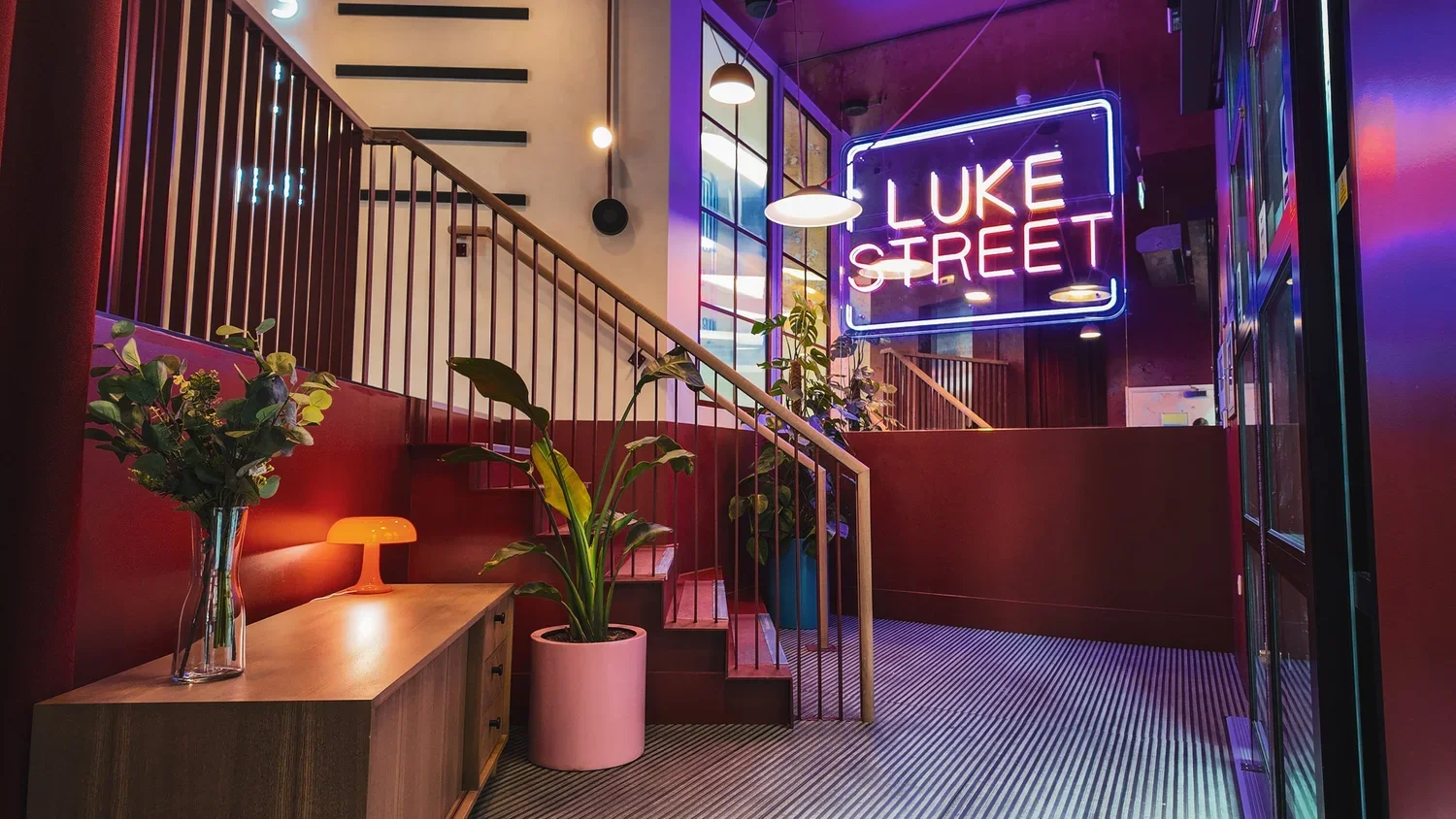 Interior of a modern lounge or bar with a neon sign that says 'LUKE STREET', a potted plant, a small lamp, and a staircase with a railing, illuminated by colorful lighting.
