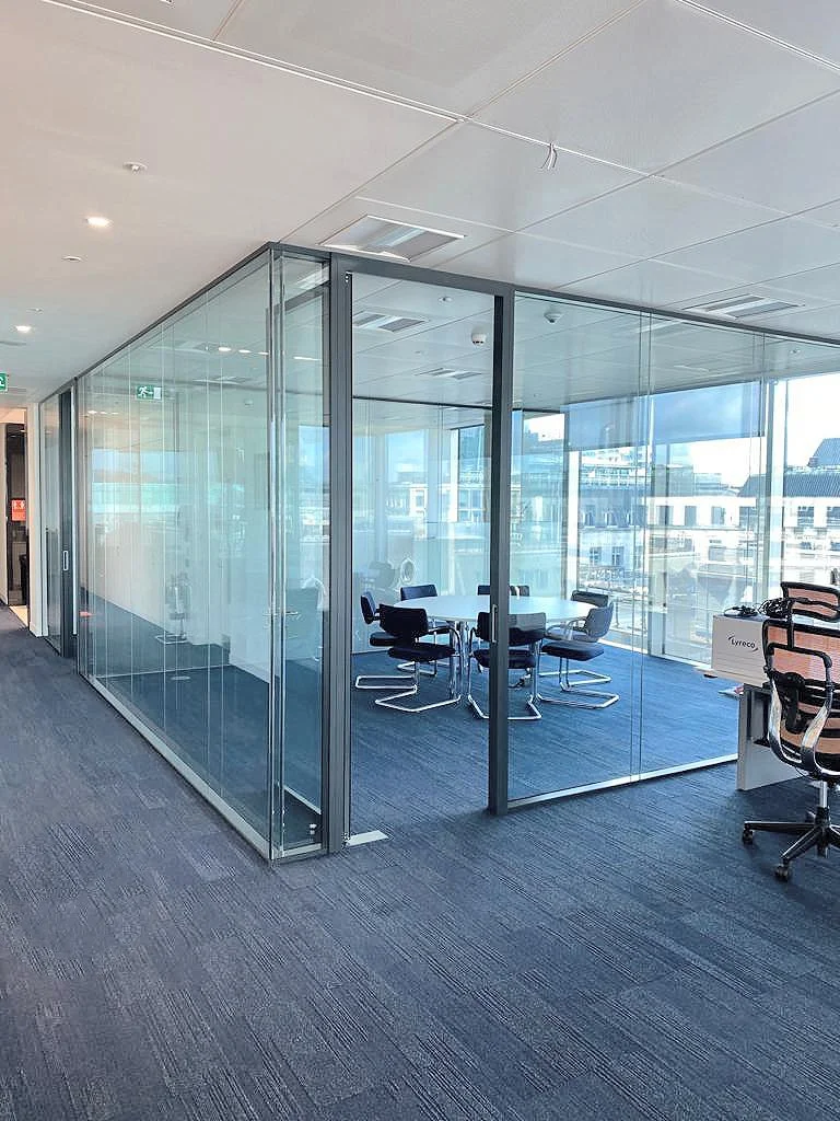Modern office conference room with glass walls, a round table, and six black chairs. Large windows with city view, gray carpet, and office furniture outside the room.