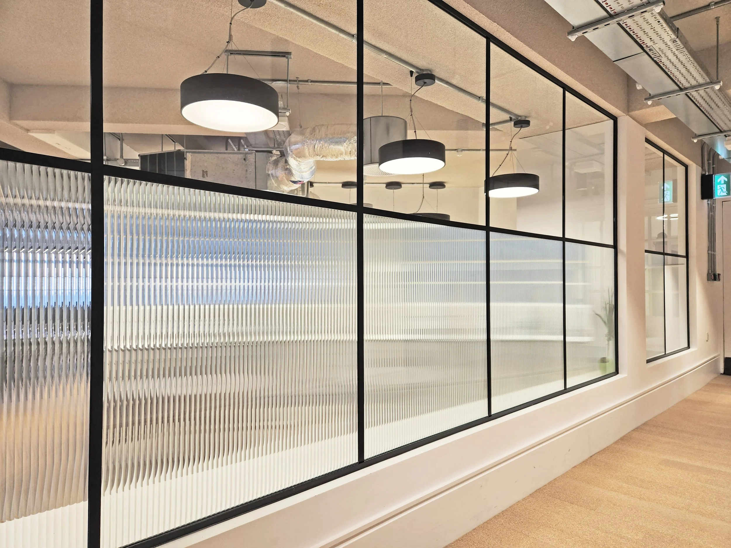 Interior view of an office or commercial space with large glass windows, modern pendant lights, exposed ductwork, and an emergency exit sign.