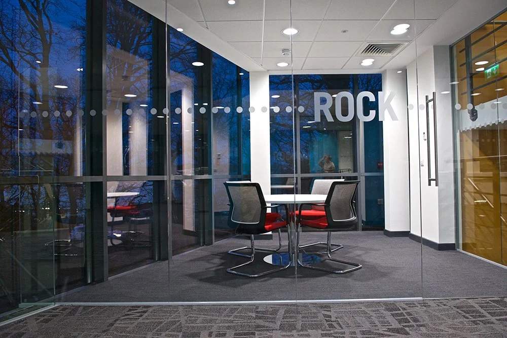 Empty modern glass meeting room with four chairs and a round table inside an office, with the word 'ROCK' on the glass window.