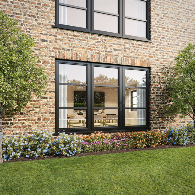 A brick house with large black-framed windows, a well-maintained lawn, and a flower bed with colorful flowers and small trees.