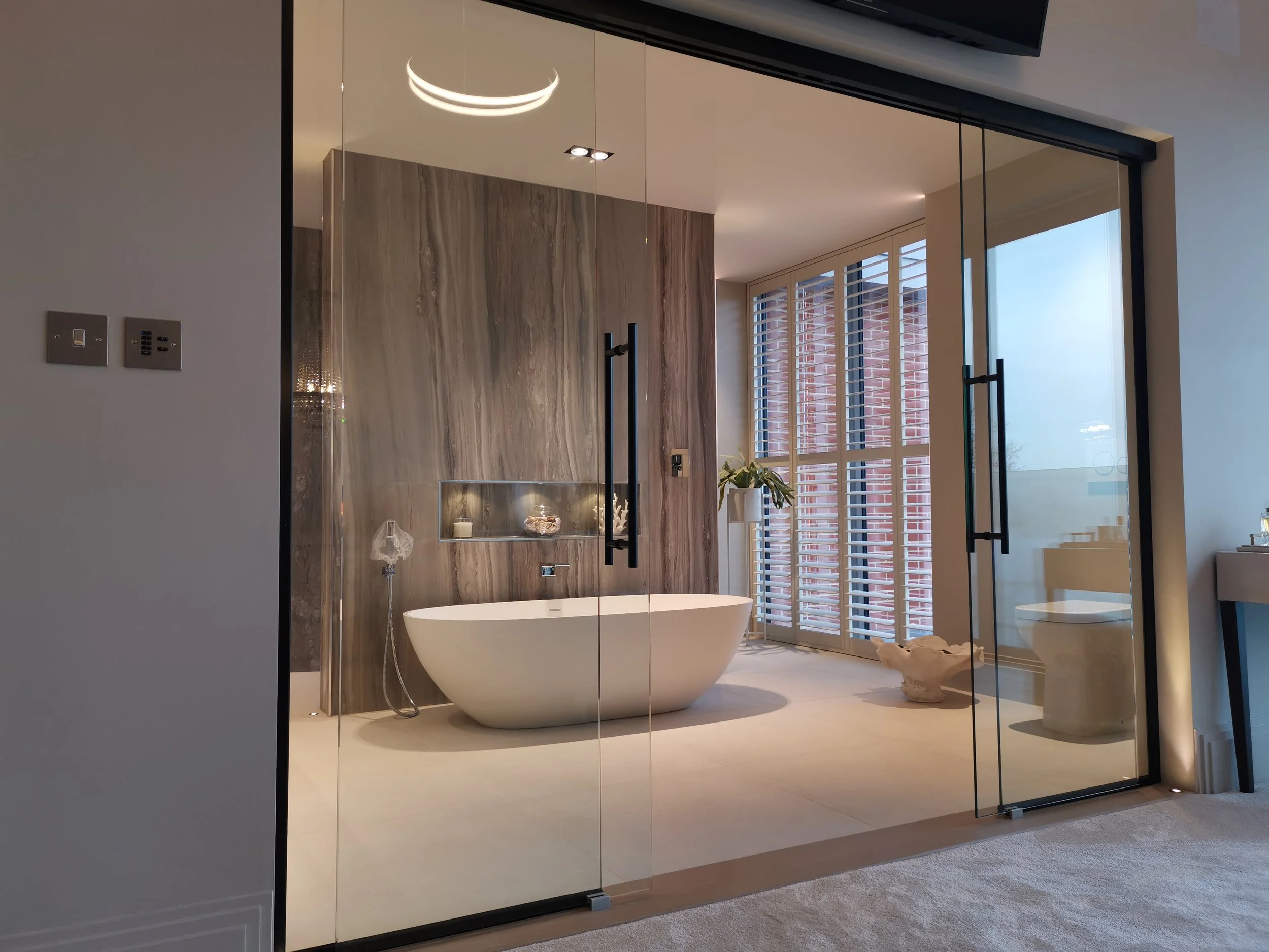 A bathroom with a freestanding bathtub, a wood-paneled accent wall, a window with white blinds, a plant on a small table, and a glass shower door.