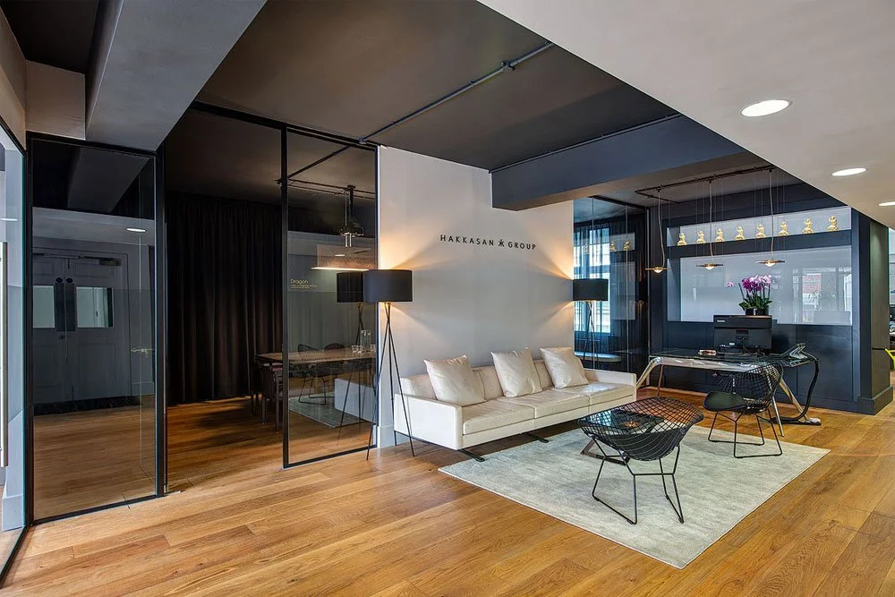 Modern office reception area with a white sofa, black wire chairs, and a gray area rug. There's a black desk with a computer and flowers, and a sign on the wall reads 'Hakkasan Group.'