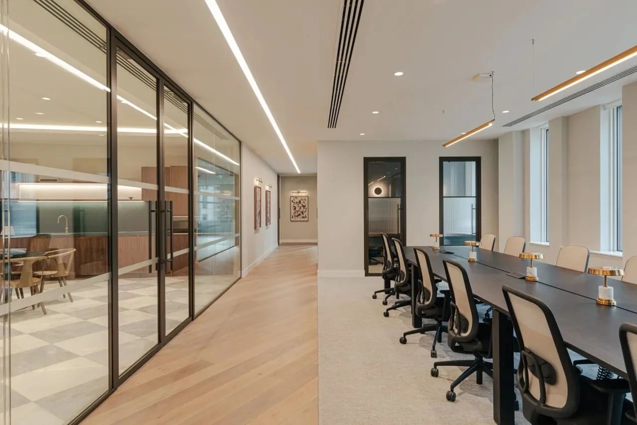 Modern conference room with a long black table, cream chairs, and large windows on the right. On the left, a glass wall separates it from another room with wooden chairs and a table. Light wood flooring and minimalistic decor.
