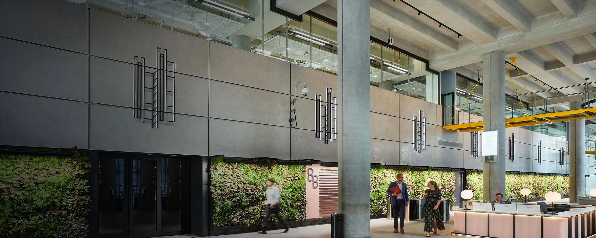 Interior of a modern building with concrete pillars, geometric wall decor, and a living green wall with people walking and talking.