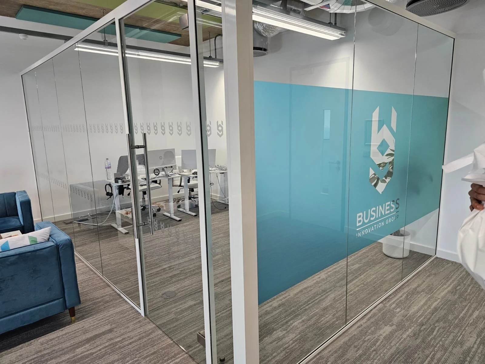 Office space enclosed by glass walls featuring company logo and name 'BUSINES S INNOVATION GROUP' on the glass. Inside, there are work desks with computers, chairs, and office supplies. The room has modern decor with light-colored walls and a gray carpet.