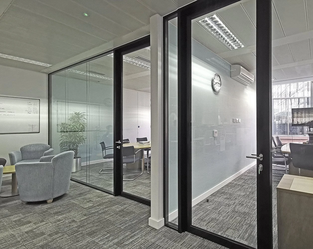 Modern office interior with a glass door, a clock on the wall, and a mix of seating including armchairs and conference chairs.
