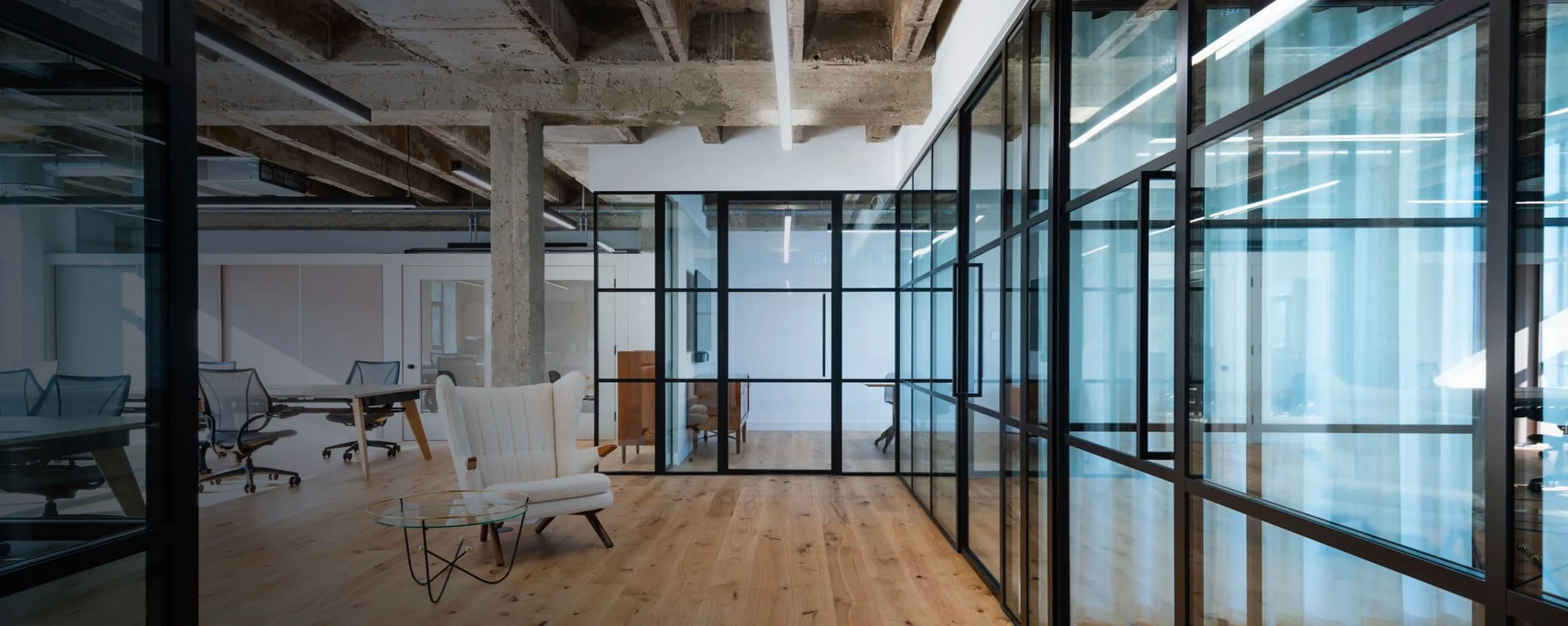 Modern office with glass partitions, wooden flooring, exposed ceiling beams, and contemporary furniture.