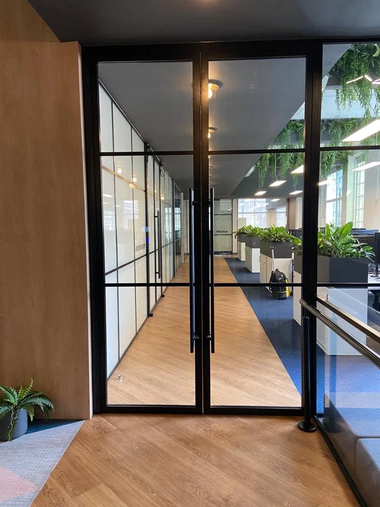 Interior view of an office space seen through black-framed glass double doors, with a wooden floor, white wall with glass panels, and large windows with greenery outside.