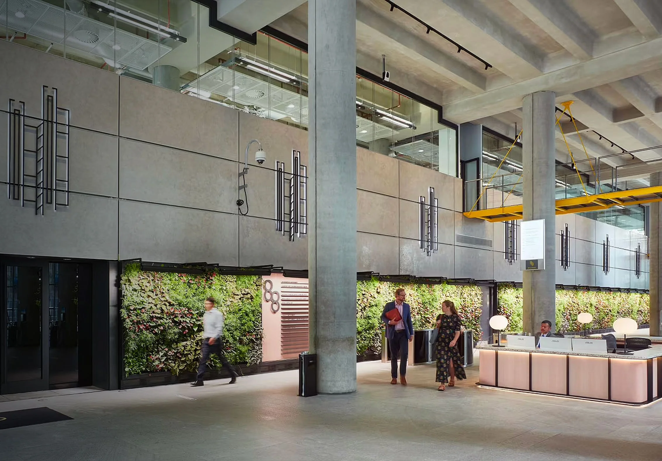 Modern interior of a building with concrete columns, glass windows, and a vertical green wall decorated with plants. People are walking and talking in the lobby area that features a reception desk and lounge seating.