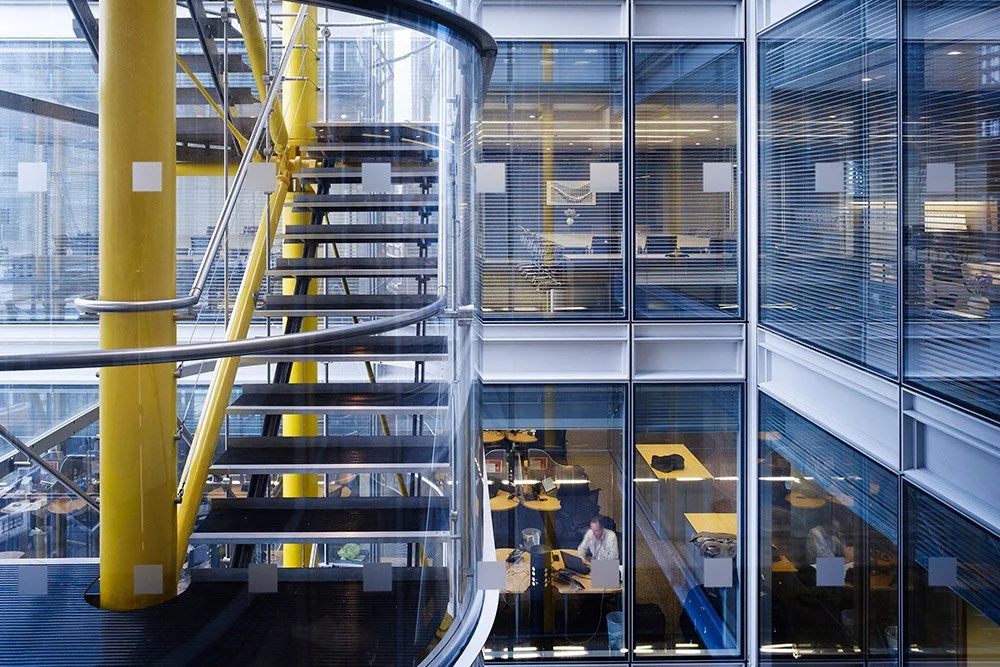 Interior view of a modern office building with glass walls, yellow structural columns, and a spiral staircase with black steps and a glass railing. Through the glass, office desks and workers are visible.