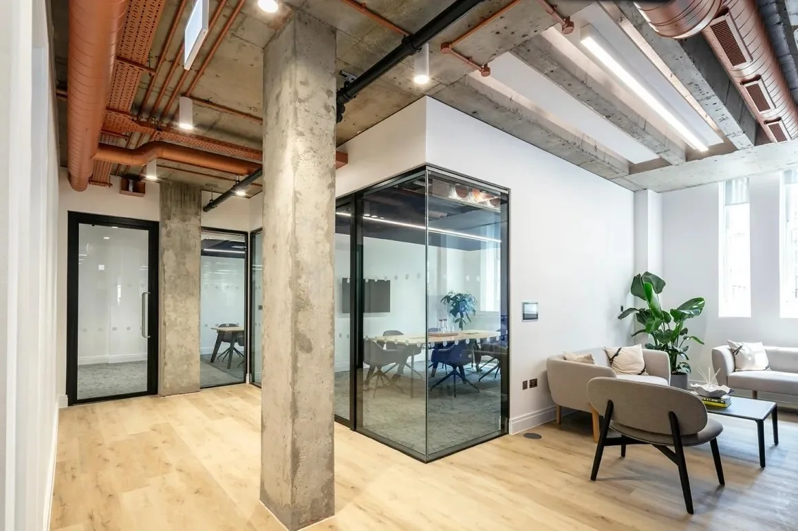 Modern office space with concrete columns, glass-walled conference room, and lounge area with white sofas and a plant.