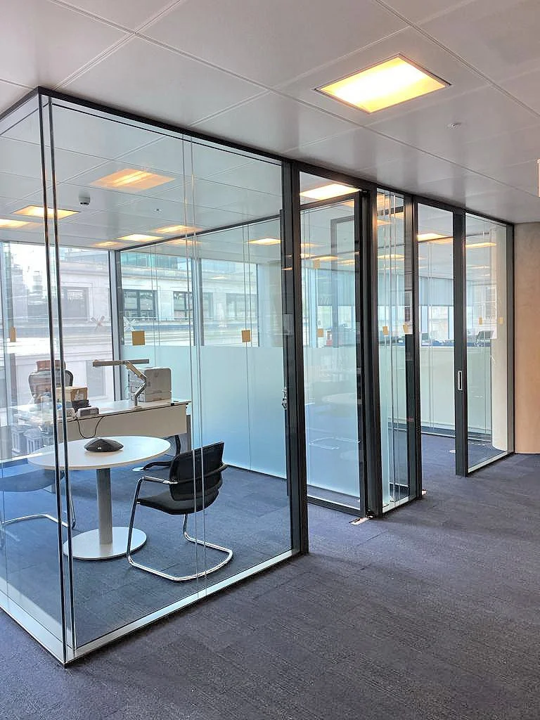 An office space with glass-walled meeting rooms, a round table, chairs, and bright ceiling lights.