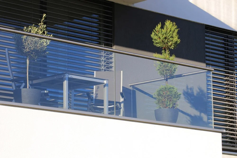 Modern balcony with glass railing, two potted trees, and black window shutters.